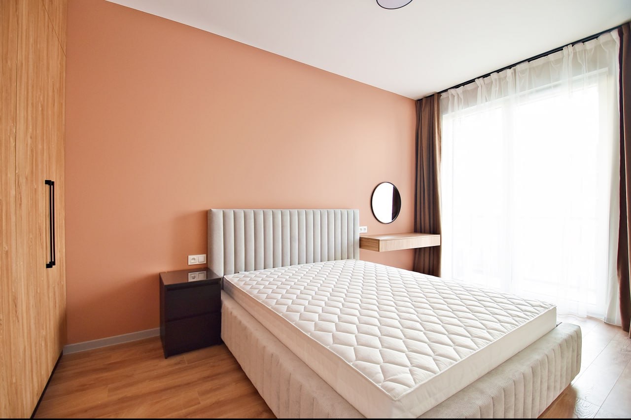 Pokój w Warszawie po odświeżeniu: łóżko z szarym zagłówkiem, ściana w odcieniach brzoskwini, drewniana szafa z czarnym uchwytem, okrągłe lustro i toaletka.