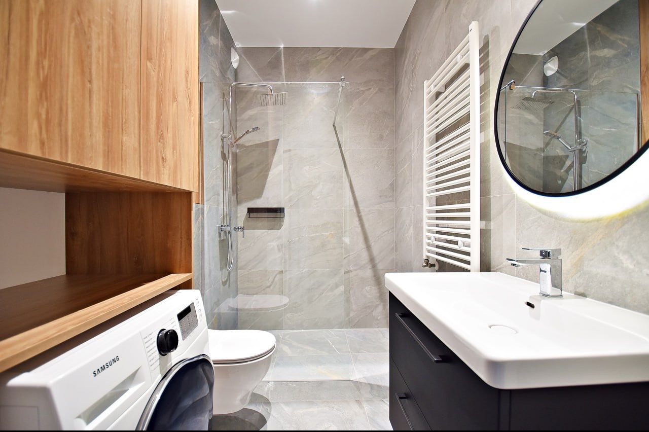 Nowoczesna łazienka z kabiną prysznicową typu walk-in, umywalką w zabudowie i pralką pod drewnianymi szafkami, wykończona szarą płytką imitującą marmur.