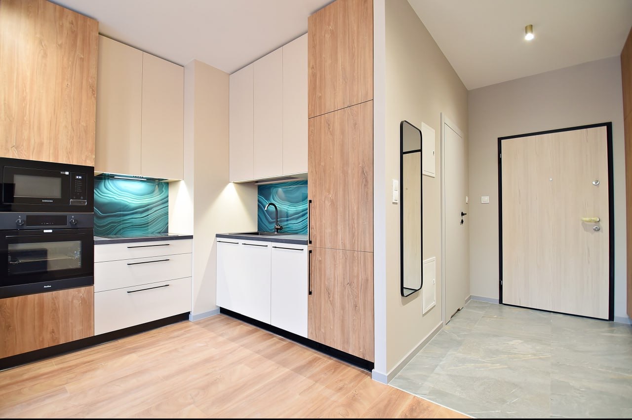 Nowoczesna kuchnia z drewnianymi i białymi szafkami, turkusowym panelem nad blatem oraz podłogą imitującą jasne drewno, widziana z perspektywy wejścia do mieszkania.