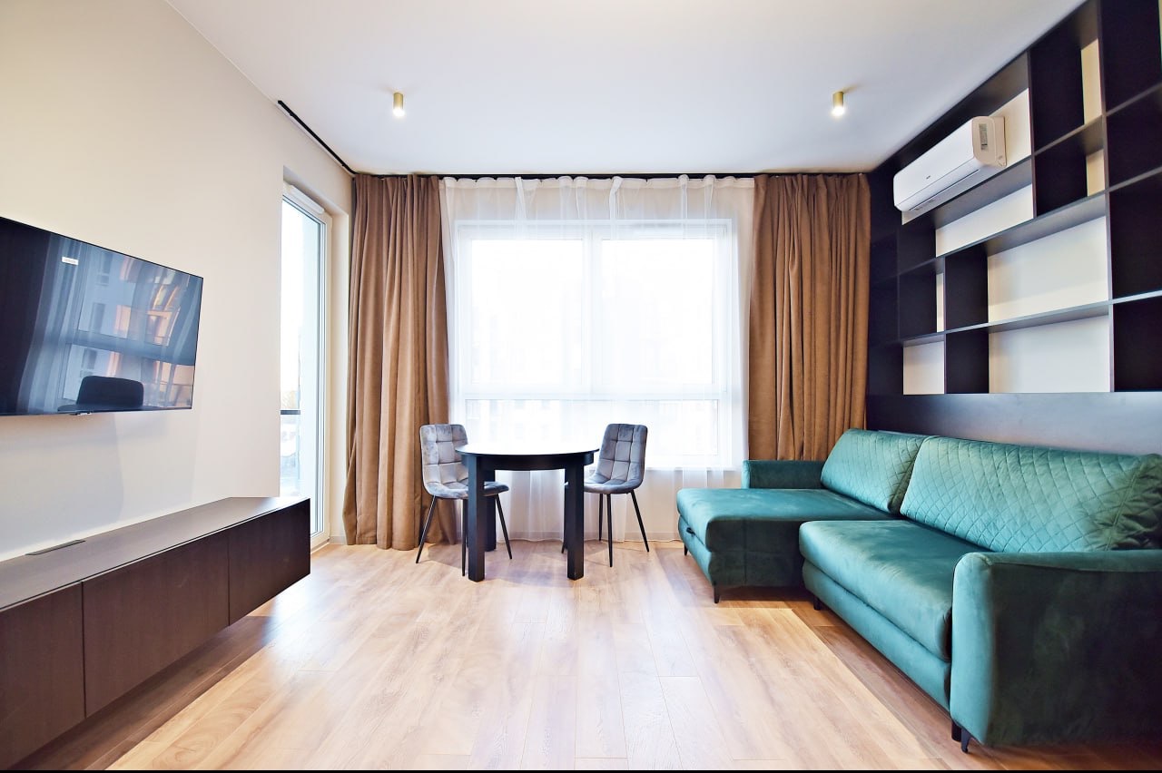 Jasne wnętrze salonu z drewnianą podłogą, zieloną sofą narożną, czarnym regałem, telewizorem na ścianie, okrągłym stołem i dwoma szarymi krzesłami w stylu glamour.