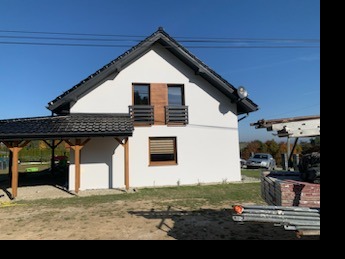 Nowoczesny dom jednorodzinny z jasną elewacją, balkonami z drewnianymi panelami i ciemnym dachem. Widoczne rusztowania i materiały budowlane sugerują prace wykończeniowe.