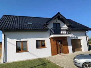 Nowoczesny dom jednorodzinny z białą elewacją, ciemnym dachem i drewnianymi elementami wykończeniowymi. Widoczny balkon z czarną balustradą i okna w kolorze drewna. Na pierwszym planie fragment...