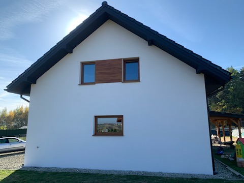 Boczny widok domu z białą, gładką elewacją, ciemnym dachem i drewnianymi akcentami okiennymi, sfotografowany w słoneczny dzień.