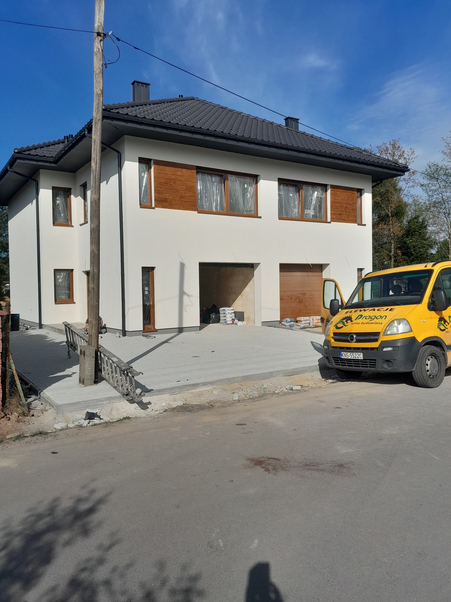 Dwukondygnacyjny dom z białą elewacją, drewnianymi akcentami okiennymi i ciemnym dachem, częściowo zakryty żółtym samochodem dostawczym firmy budowlanej, z workami materiałów budowlanych widocznymi...