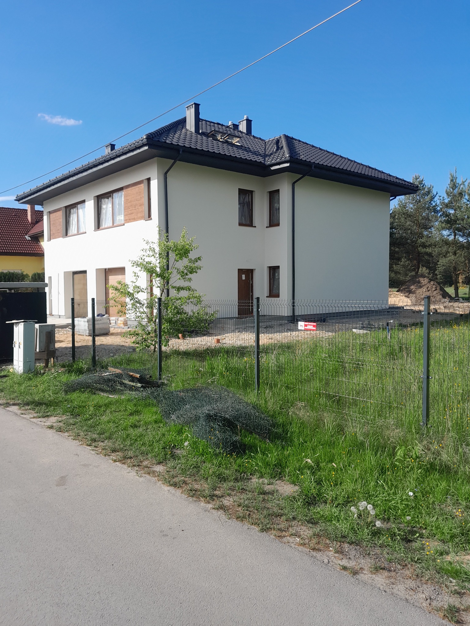 Dwupiętrowy dom jednorodzinny z jasną elewacją i ciemnym dachem, ogrodzony metalową siatką, widok z poziomu ulicy, teren jeszcze w trakcie zagospodarowywania.