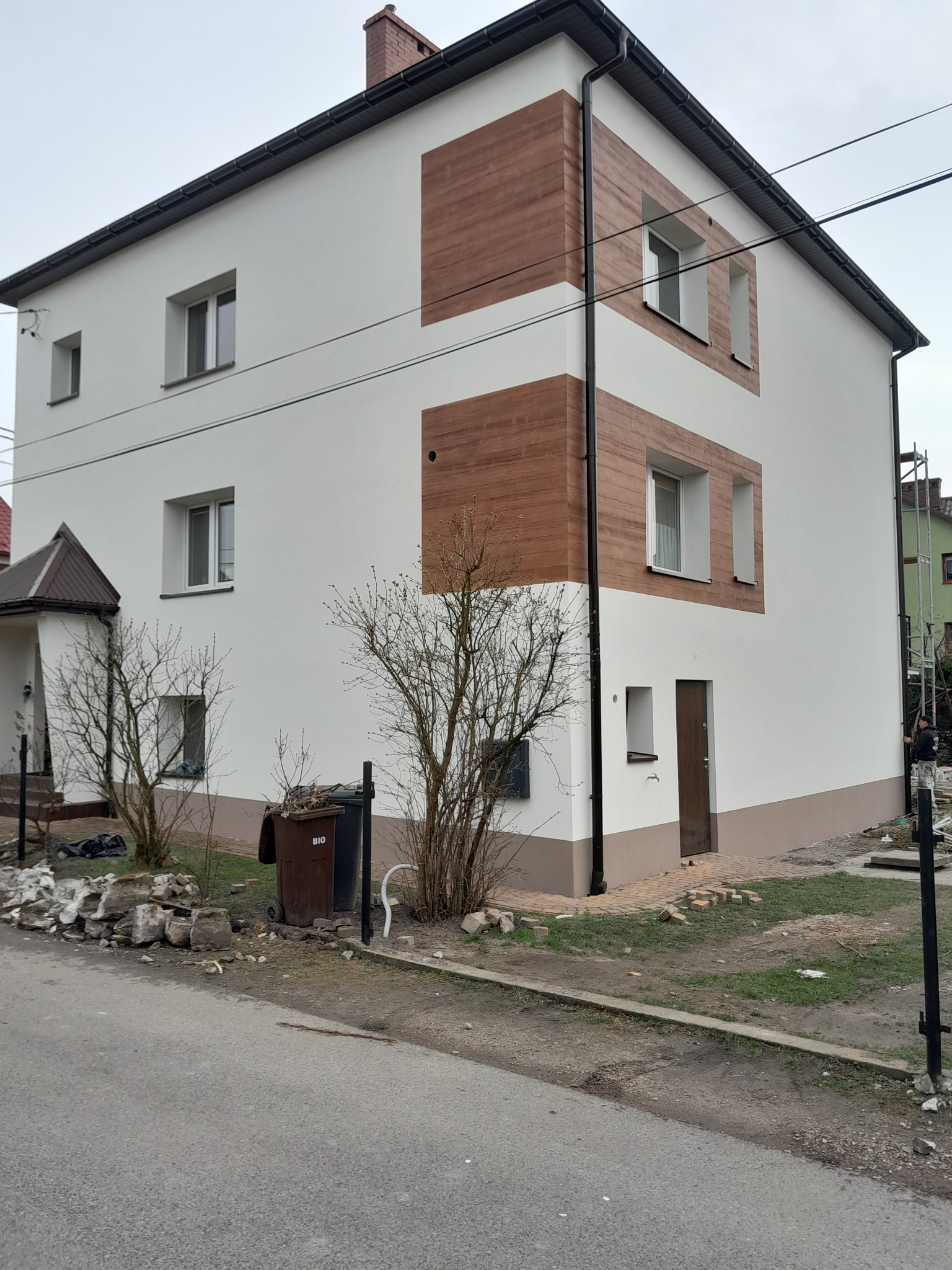 Dwukondygnacyjny dom z białą elewacją i dekoracyjnymi panelami imitującymi drewno, widoczne ślady prac wykończeniowych na posesji.