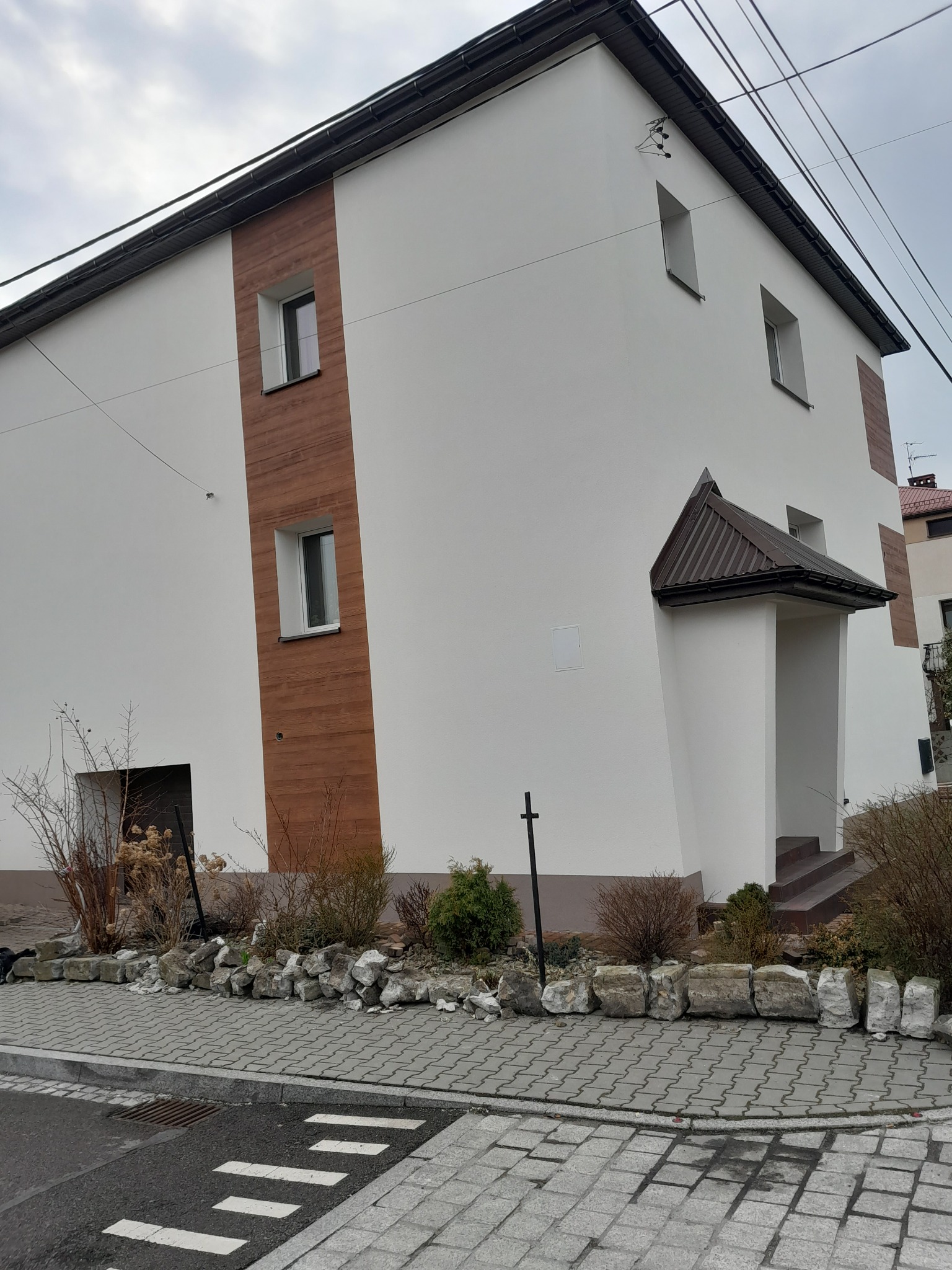 Dwukondygnacyjny dom z białymi ścianami i drewnopodobnymi panelami elewacyjnymi, widok z zewnątrz na elewację frontową i boczną, z wejściem zadaszonym brązowym daszkiem.