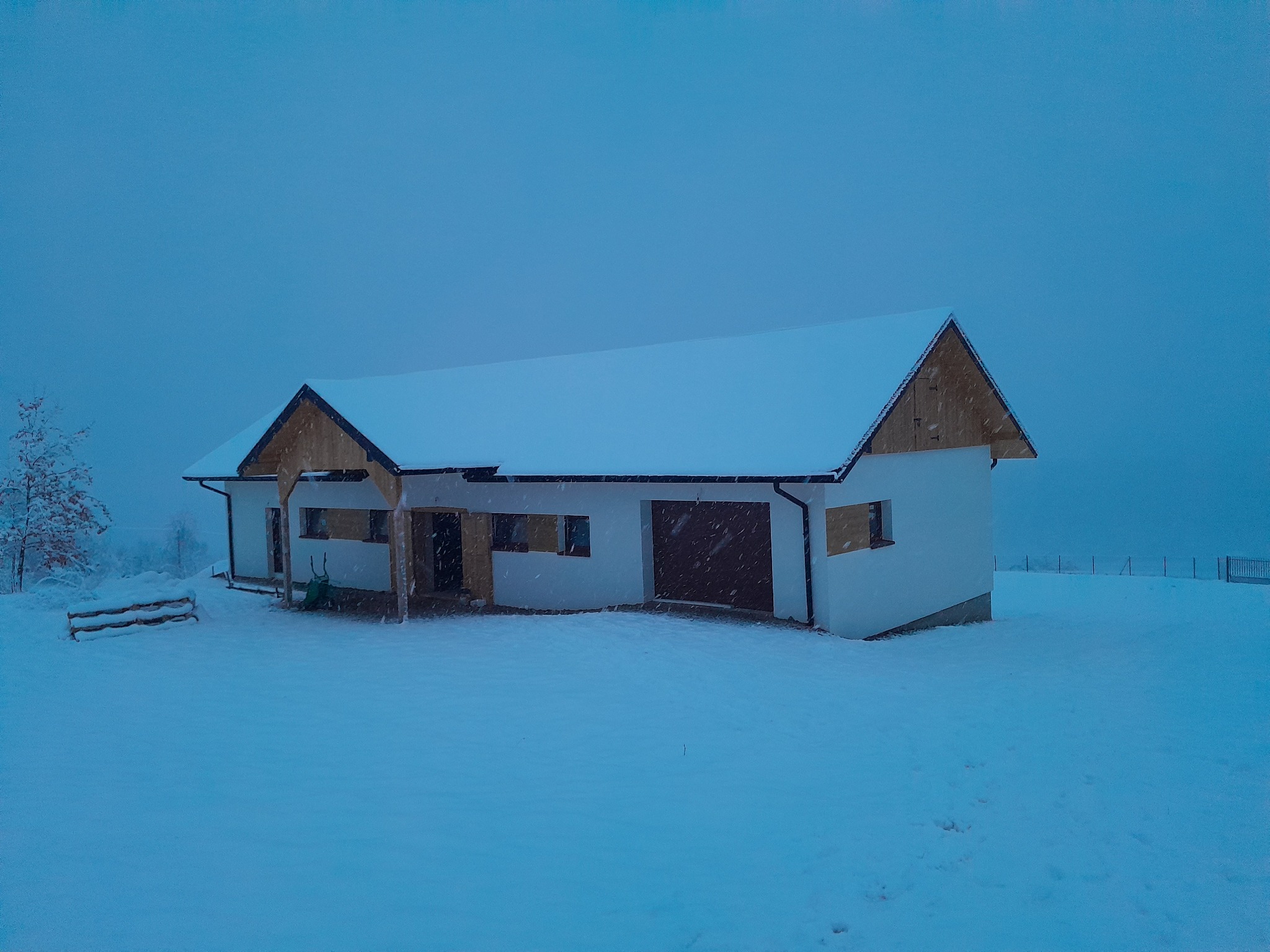 Nowo wybudowany, biały dom jednorodzinny z brązową bramą garażową, przysypany śniegiem w zimowej scenerii. Widoczne ośnieżone drzewa w tle i padający śnieg.