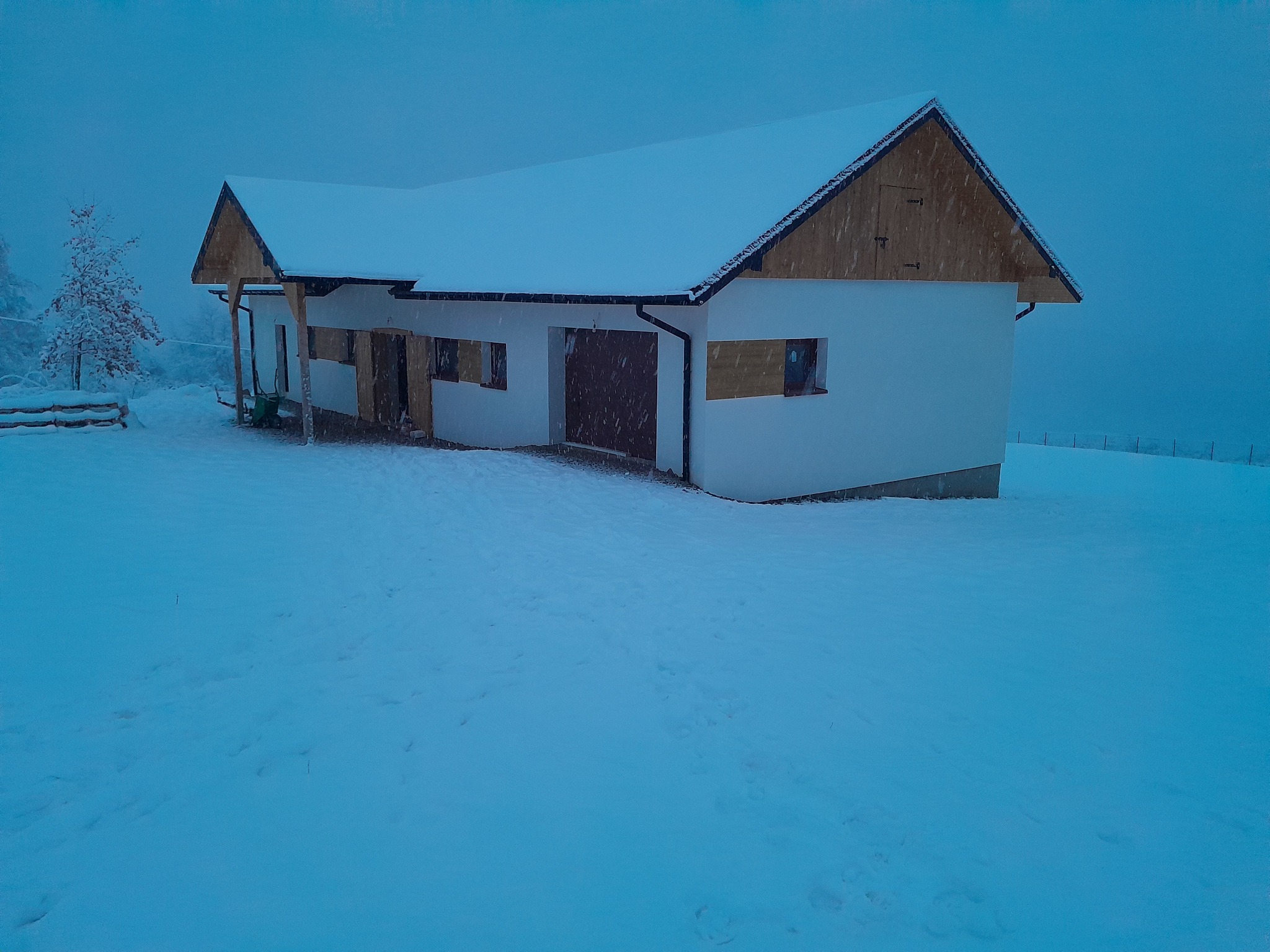 Świeżo otynkowany, biały budynek gospodarczy z drewnianym szczytem dachu, pokryty śniegiem podczas zimowej zamieci.