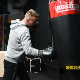 Mężczyzna w szarej bluzie z kapturem i czarnych rękawicach bokserskich Daniken uderza w czarny worek treningowy Bushido w ciemnym pomieszczeniu.