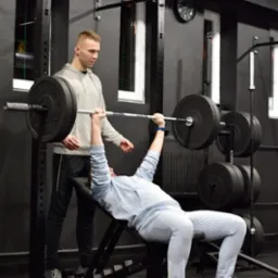 Mężczyzna ćwiczy wyciskanie sztangi na ławce pod okiem instruktora w siłowni z czarnymi ścianami i sprzętem.