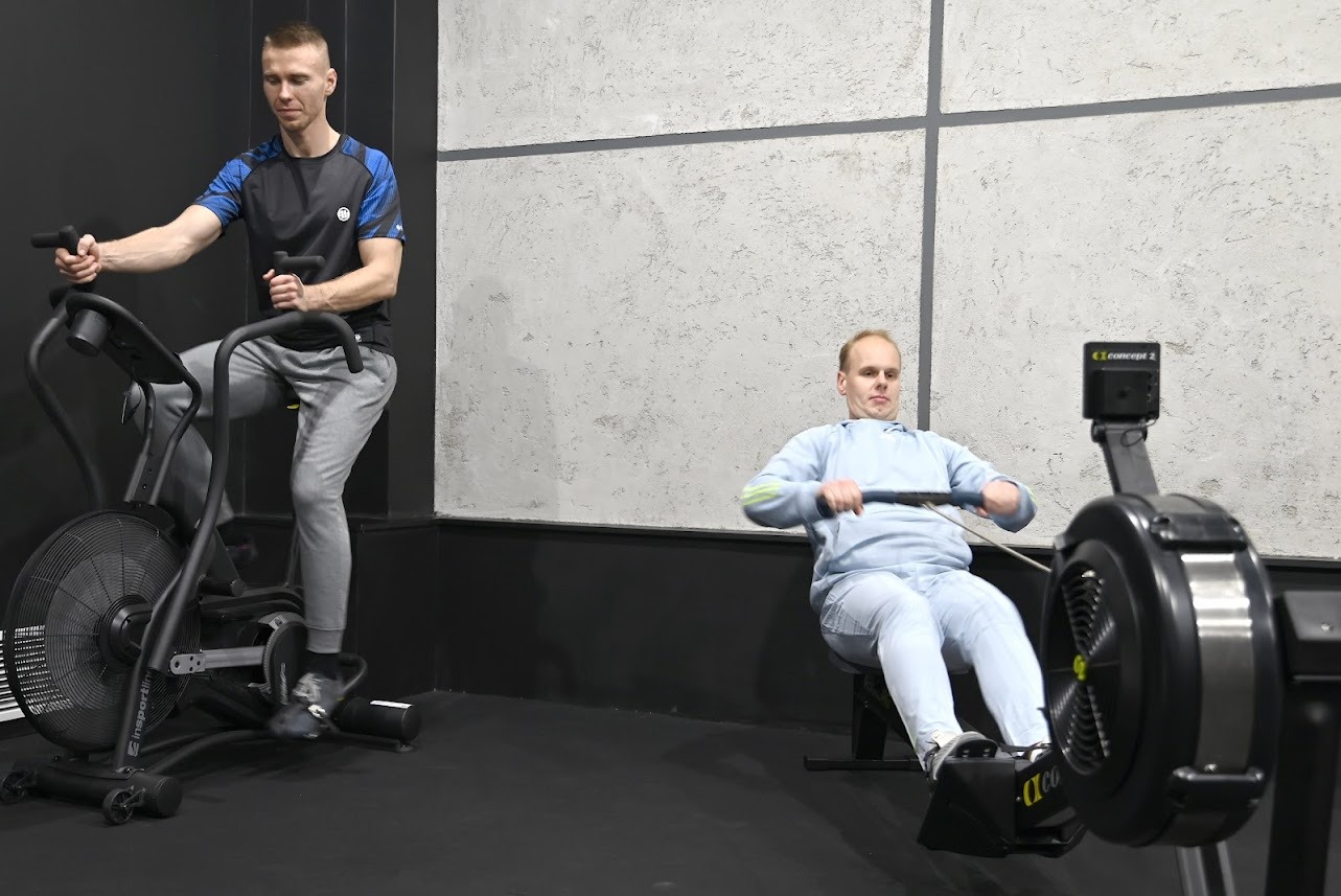 Dwaj mężczyźni ćwiczą na sprzęcie fitness: jeden na rowerze powietrznym Insportline, drugi na ergometrze wioślarskim Concept 2, w siłowni z szarą ścianą w tle.