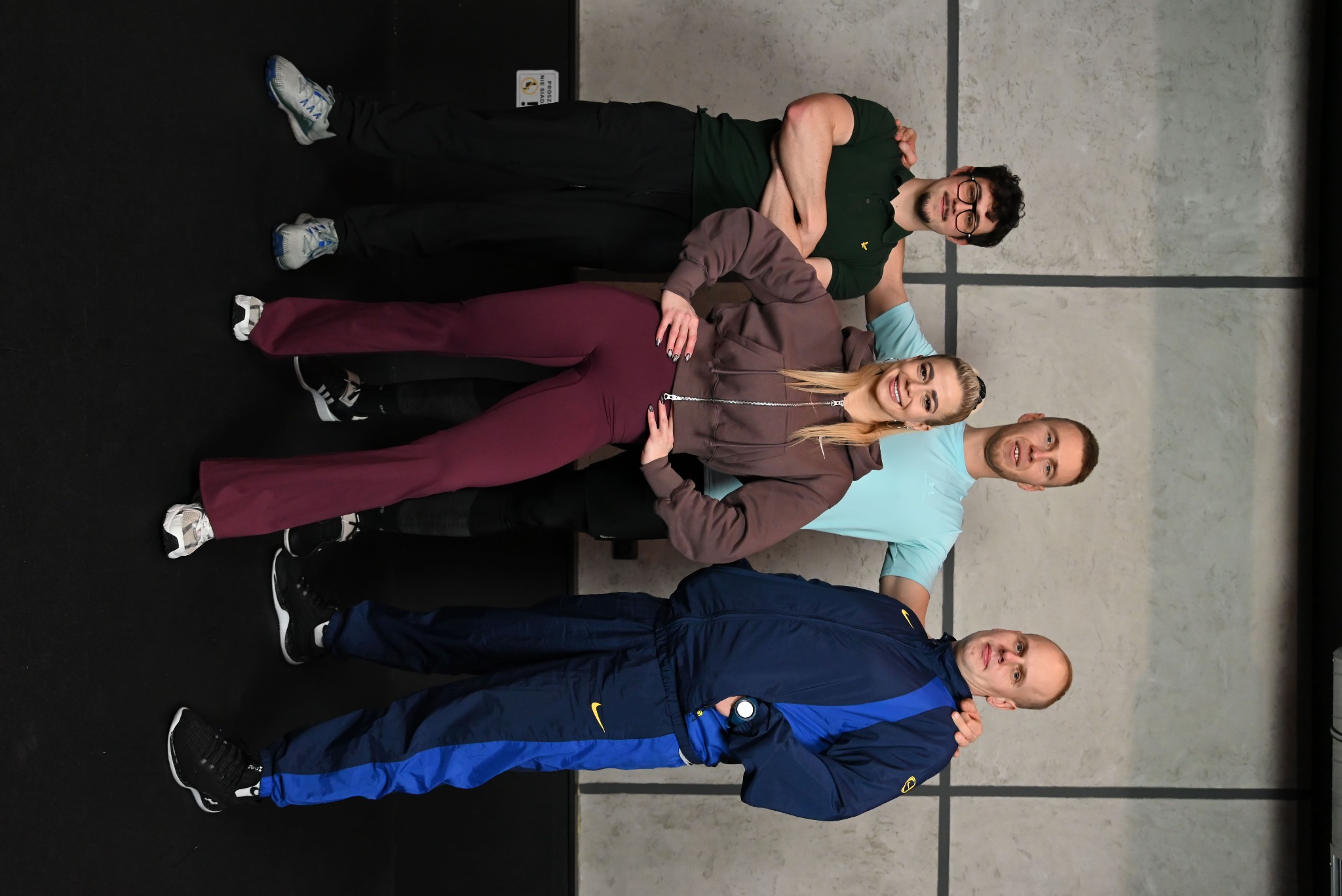 Cztery osoby w sportowych strojach leżące obok siebie na podłodze w siłowni, uśmiechnięte, w nietypowej pozie. Widoczne buty sportowe i fragment ściany.