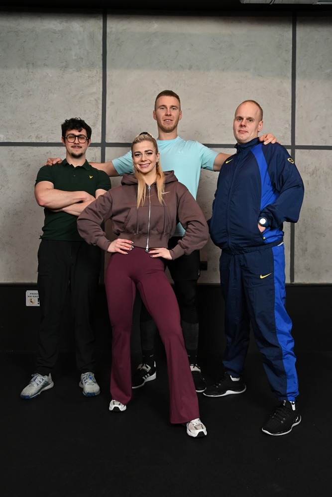 Grupa czterech osób w strojach sportowych pozuje na tle ściany w siłowni. Kobieta w centrum, z rękami na biodrach, uśmiecha się do kamery. Mężczyźni w tle i po bokach.