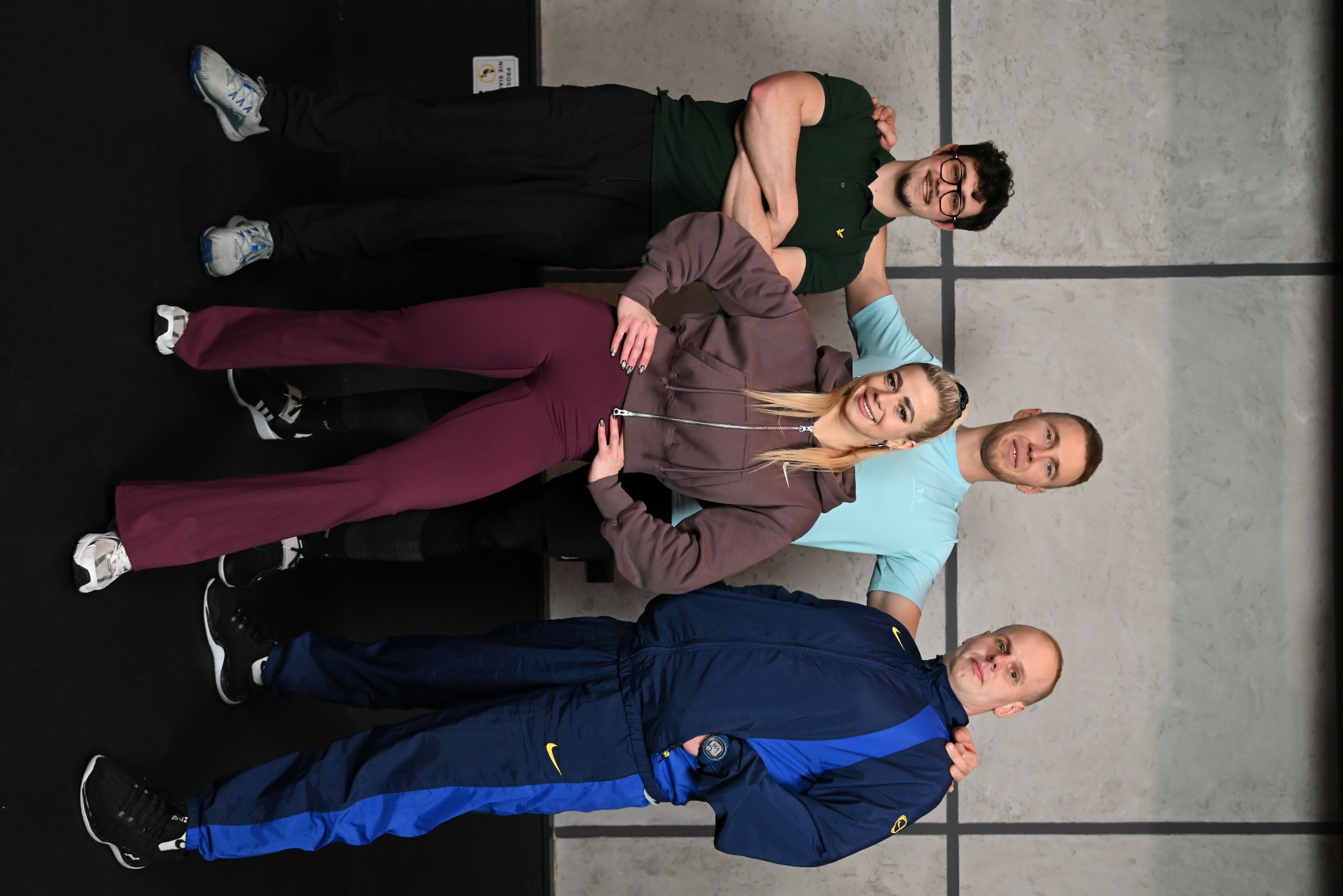 Cztery osoby w strojach sportowych, leżące obok siebie na podłodze w siłowni, uśmiechnięte, z rękami na ramionach sąsiada. Wspólny trening w zespole.