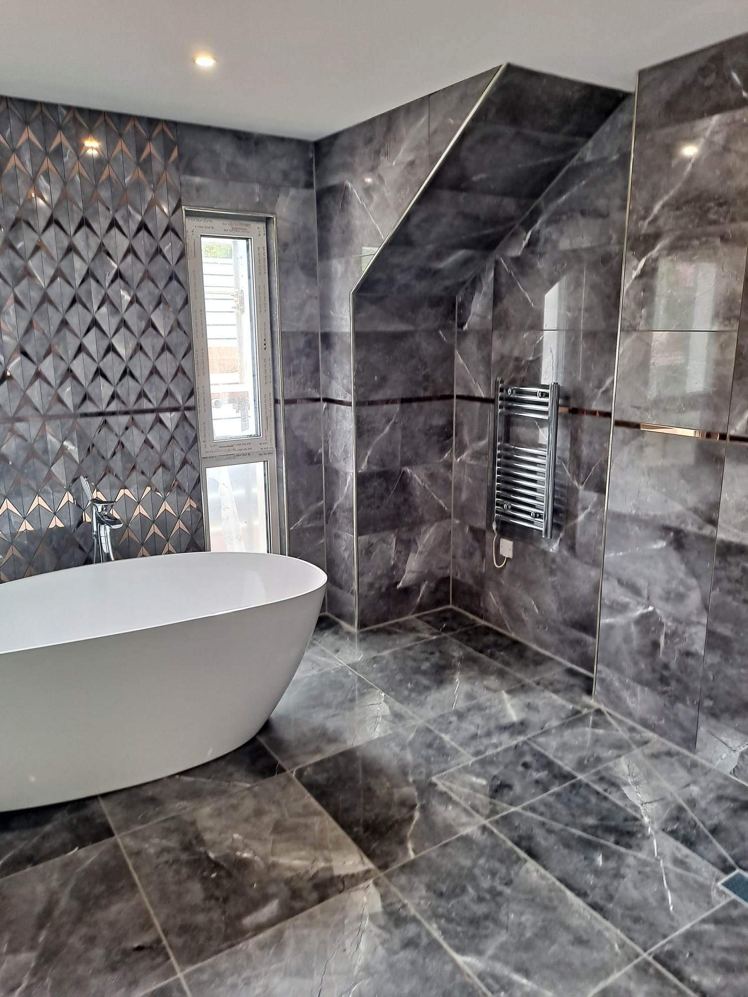 Nowoczesna łazienka z wolnostojącą wanną, szarymi marmurowymi płytkami na ścianach i podłodze, dekoracyjną ścianą z trójwymiarowych płytek i grzejnikiem drabinkowym.