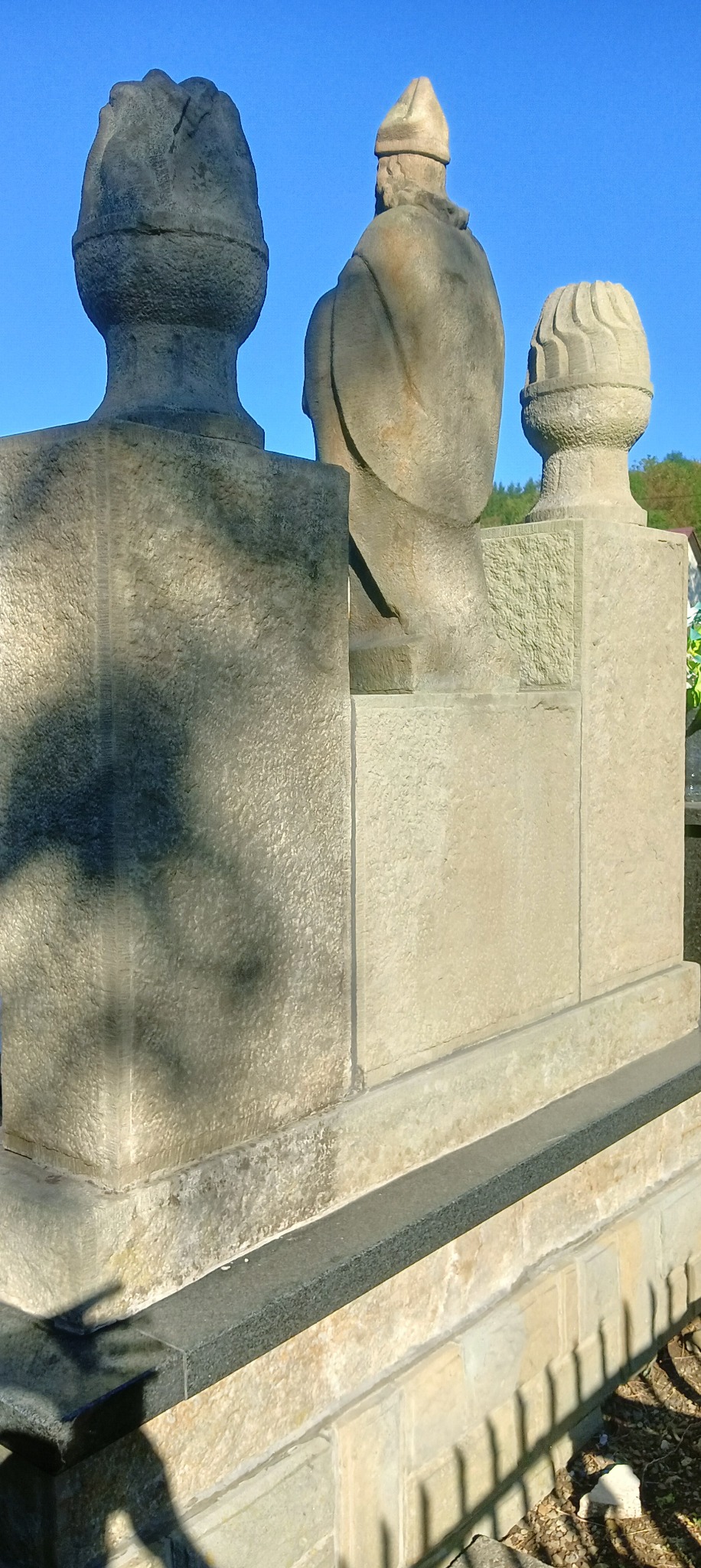 Fragment pomnika nagrobnego z trzema rzeźbami z jasnego kamienia na tle błękitnego nieba, widoczne detale obróbki kamienia i cieniowanie