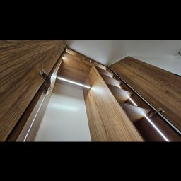 MB - meble - Wnętrze szafy z drewnianymi półkami i oświetleniem LED, widok z dołu, ukazujący precyzję wykonania i nowoczesny design.