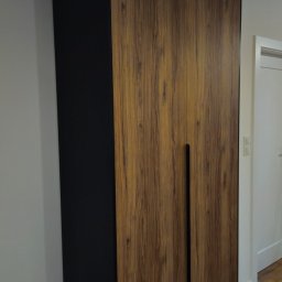 MB - meble - Dwudrzwiowa szafa z okleiną drewnopodobną i czarnymi bokami, zintegrowane uchwyty, na tle parkietu i białej ściany z gniazdkiem.