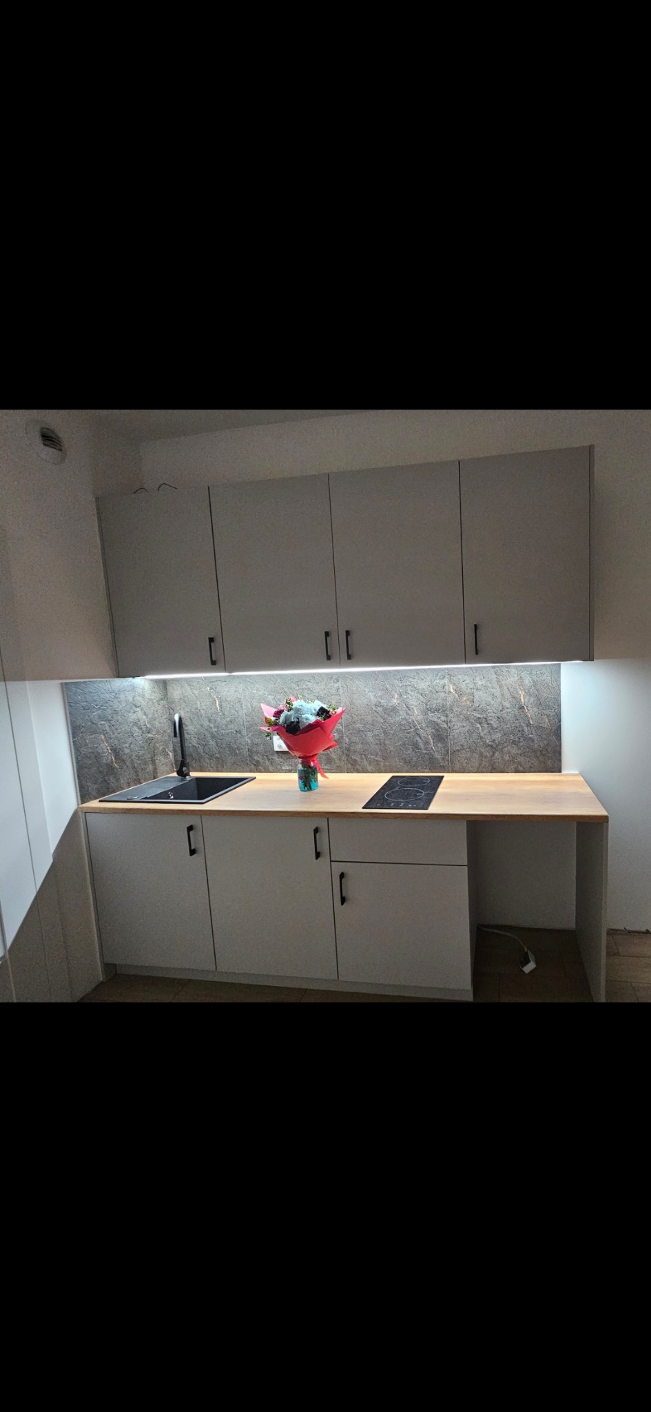 Jasna kuchnia z szarymi szafkami i drewnianym blatem, oświetlona listwą LED. Na blacie stoi bukiet kwiatów, a w tle widoczna marmurowa ściana.