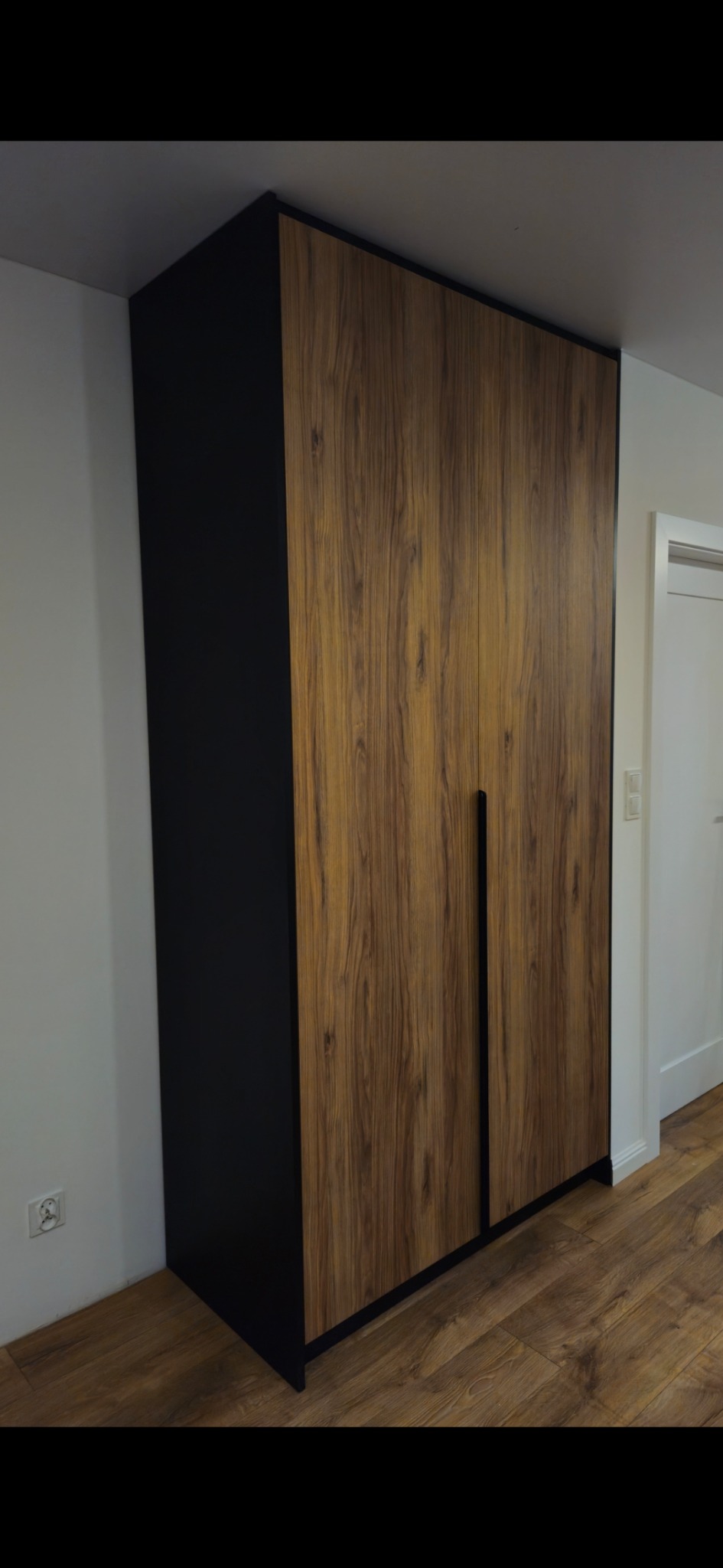 Dwudrzwiowa szafa z okleiną drewnopodobną i czarnymi bokami, zintegrowane uchwyty, na tle parkietu i białej ściany z gniazdkiem.