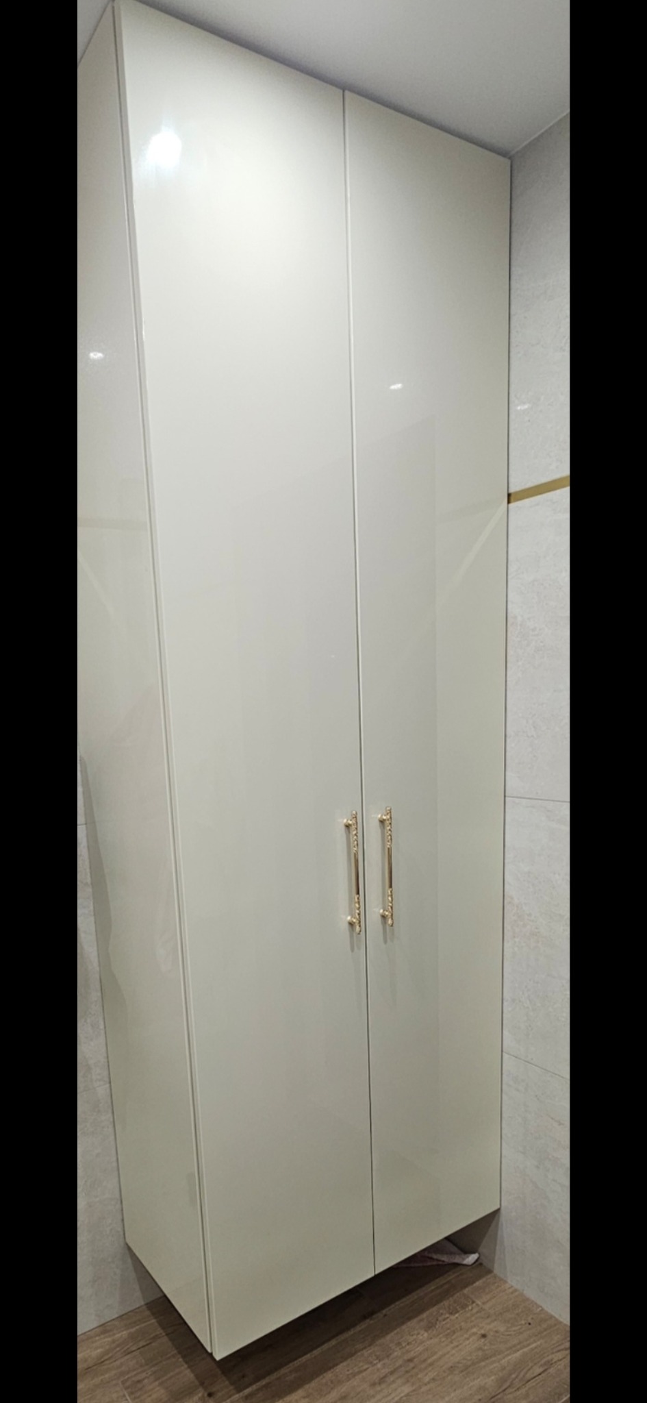 Biała, lakierowana szafa wnękowa z dekoracyjnymi, złotymi uchwytami na tle jasnoszarych płytek i drewnopodobnej podłogi. Minimalistyczny design.