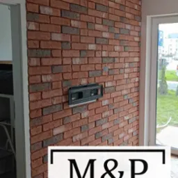 Ściana wykończona czerwoną cegłą klinkierową z widoczną puszką instalacyjną pod telewizor, obok okno z widokiem na zewnątrz.