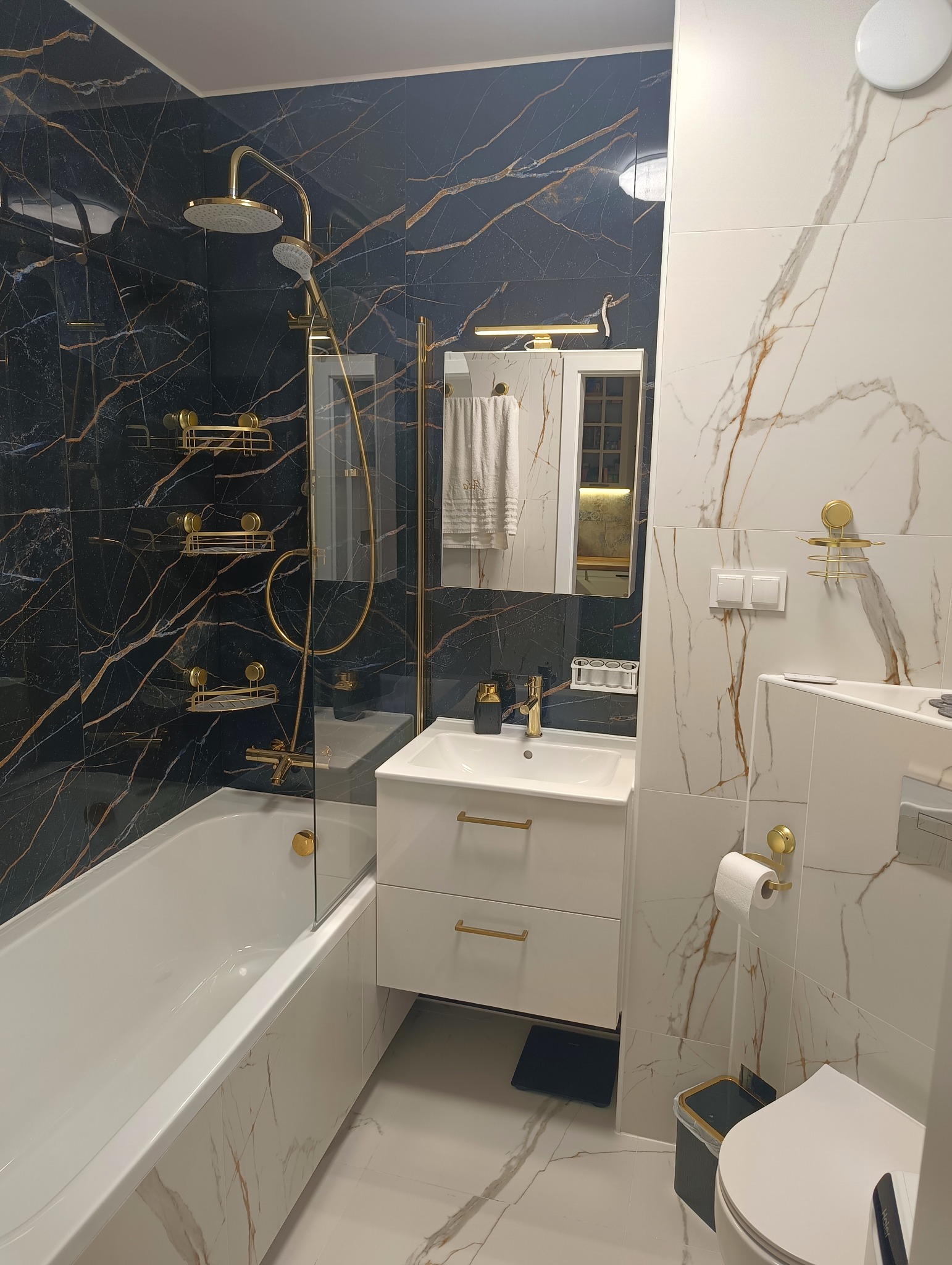 Nowoczesna łazienka z czarnymi i białymi marmurkowymi płytkami, złotą armaturą i minimalistyczną białą szafką pod umywalką, widok z poziomu podłogi.