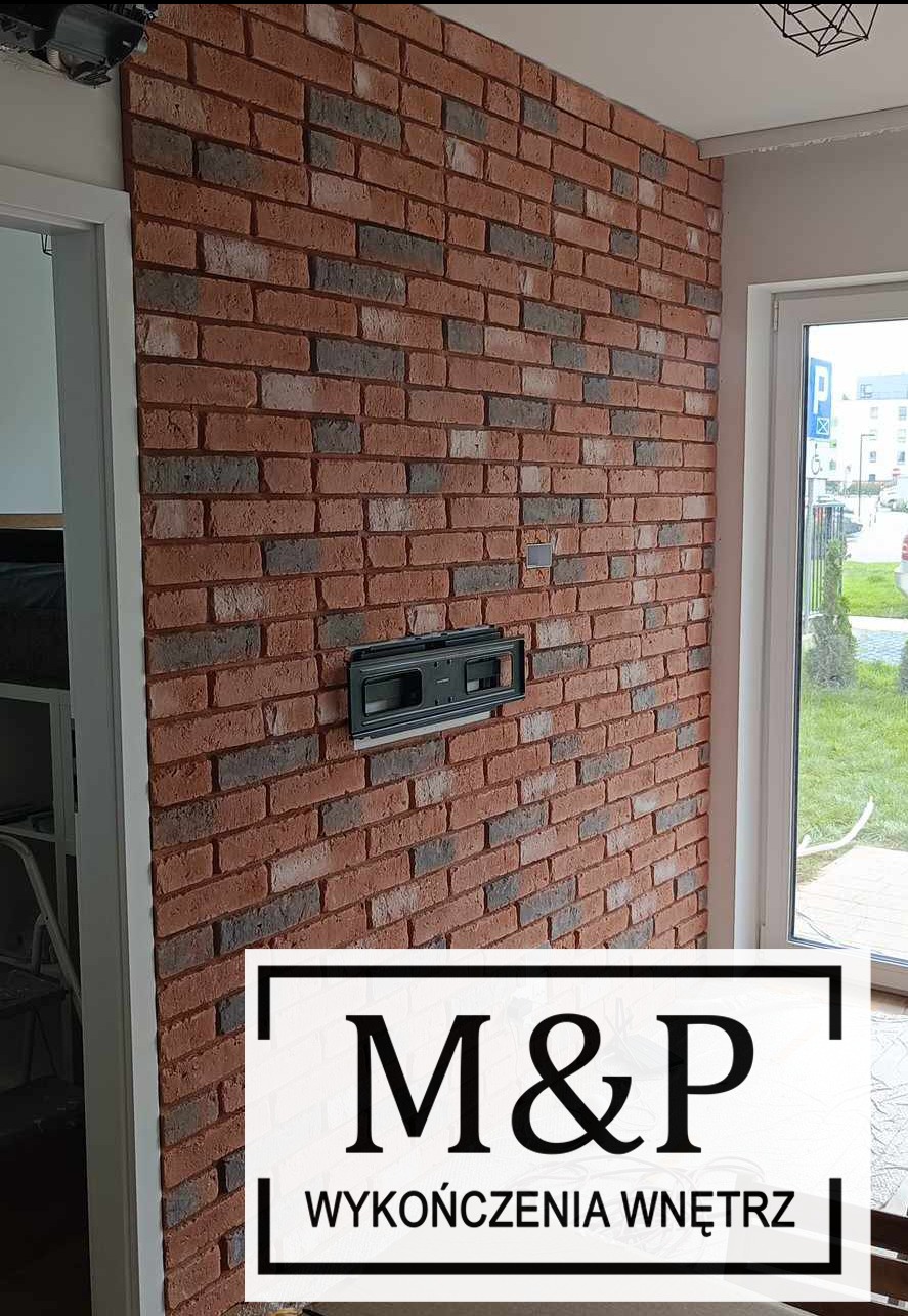 Ściana wykończona czerwoną cegłą klinkierową z widoczną puszką instalacyjną pod telewizor, obok okno z widokiem na zewnątrz.