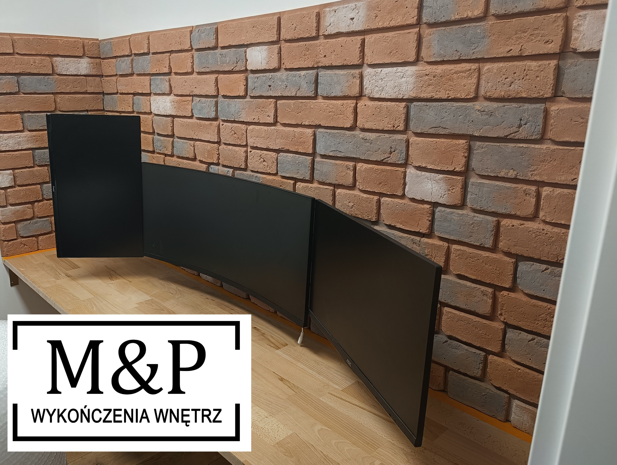 Ściana z cegły dekoracyjnej w odcieniach brązu i szarości, za trzema czarnymi monitorami na drewnianym blacie; widoczny fragment logo firmy 'M&P Wykończenia Wnętrz'.