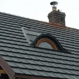 Ciemnoszara dachówka modułowa na skośnym dachu z oknem typu wole oko i ceglanym kominem z metalowym daszkiem.