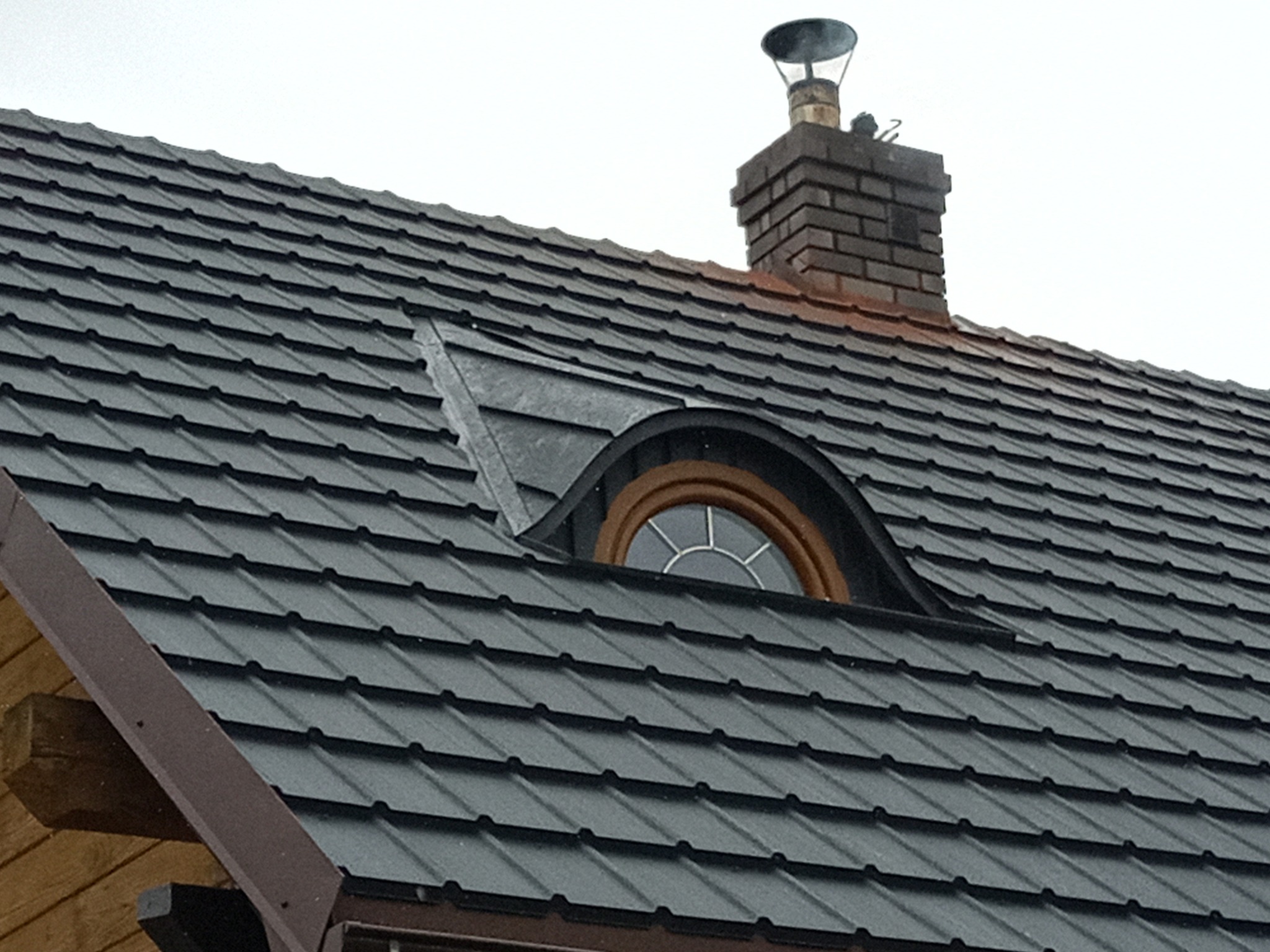 Ciemnoszara dachówka modułowa na skośnym dachu z oknem typu wole oko i ceglanym kominem z metalowym daszkiem.