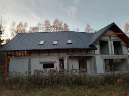 Surowy budynek w trakcie budowy z nowym, grafitowym dachem z blachy trapezowej i trzema oknami dachowymi, widok z zewnątrz na tle drzew.