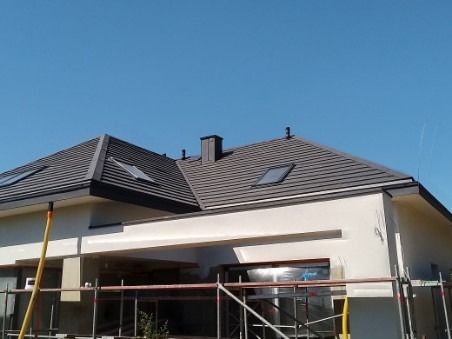 Nowoczesny dom jednorodzinny z grafitowym dachem z dachówki ceramicznej, widoczne okna dachowe i komin, elewacja w trakcie wykończenia, rusztowania przy ścianach.