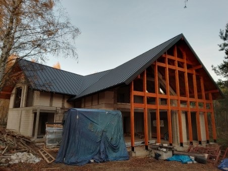 Dom w trakcie budowy z drewnianą konstrukcją szkieletową i częściowo pokrytym ciemnoszarym dachem, zabezpieczony plandeką.