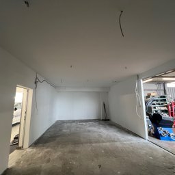 Dawid Kliczkowski - Wnętrze pomieszczenia w trakcie remontu, ściany świeżo pomalowane na biało, widoczne wystające przewody elektryczne, podłoga surowa, przez otwarte drzwi widoczny sprzęt warsztatowy.