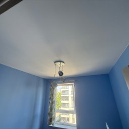 Dawid Kliczkowski - Błękitne ściany i biały sufit w pokoju z oknem. Widoczna lampa sufitowa, fragment drzwi i okno z widokiem na budynek.
