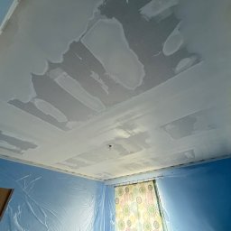 Dawid Kliczkowski - Nierównomiernie nałożona gładź na suficie z widocznymi łatami, zabezpieczona folią ściana w kolorze niebieskim i zasłonięte okno.