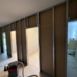 Montaż ścian działowych z płyty karton-gips na metalowym stelażu w trakcie prac wykończeniowych. Widoczny odkurzacz przemysłowy i fragment podłogi z płytek.