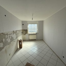 Dawid Kliczkowski - Wnętrze pokoju w trakcie remontu: częściowo usunięte stare płytki, ściany przygotowane do gładzi, okno zabezpieczone folią, na podłodze białe płytki.