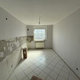 Wnętrze pokoju w trakcie remontu: częściowo usunięte stare płytki, ściany przygotowane do gładzi, okno zabezpieczone folią, na podłodze białe płytki.