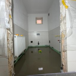 Dawid Kliczkowski - Wnętrze łazienki w trakcie wylewki samopoziomującej. Widoczne białe płytki na ścianach, grzejnik i przygotowane podłoże pod wylewkę. Folia ochronna na framugach.