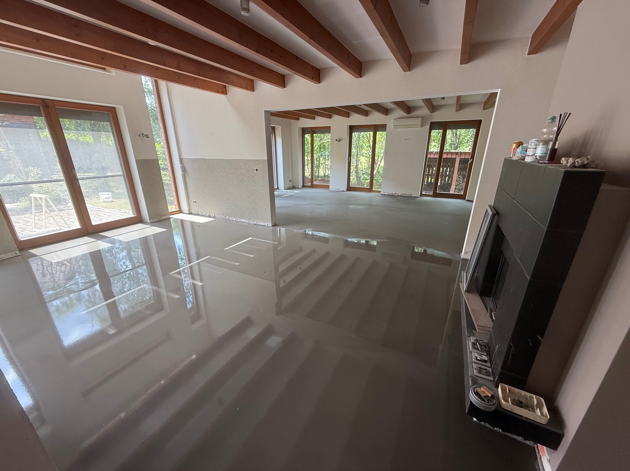 Wnętrze po wylaniu samopoziomującej wylewki, widoczne drewniane belki stropowe i duże okna. Ściany częściowo przygotowane do dalszych prac wykończeniowych.