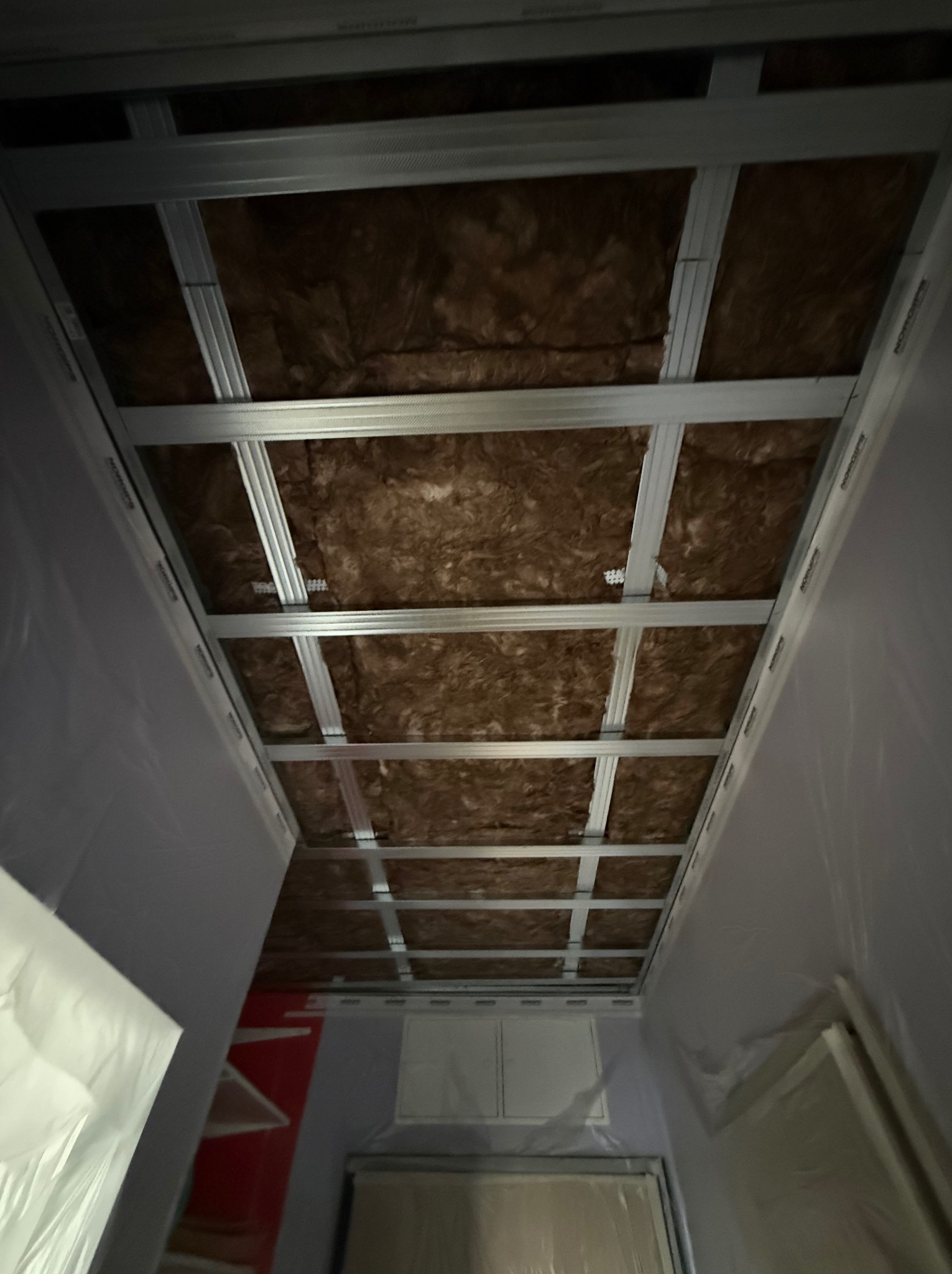 Sufit podwieszany: metalowa konstrukcja z izolacją z wełny mineralnej, widok z dołu. Ściany zabezpieczone folią malarską. Etap prac wykończeniowych.