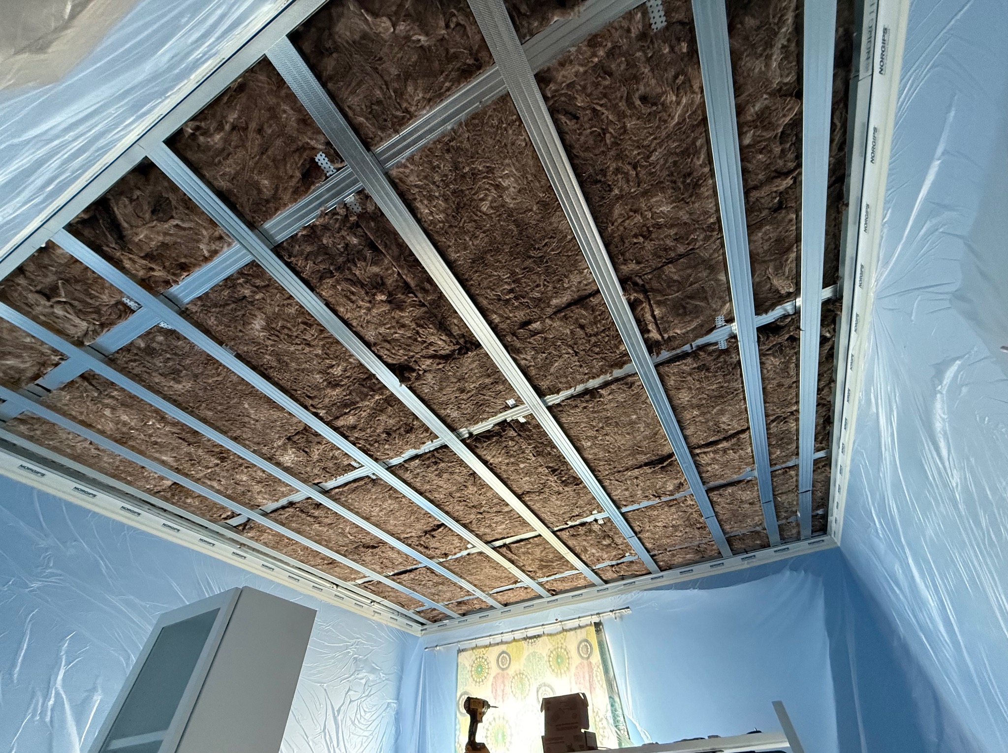 Montaż izolacji z wełny mineralnej i metalowego stelażu sufitu podwieszanego w pokoju zabezpieczonym folią malarską, widok z dołu.