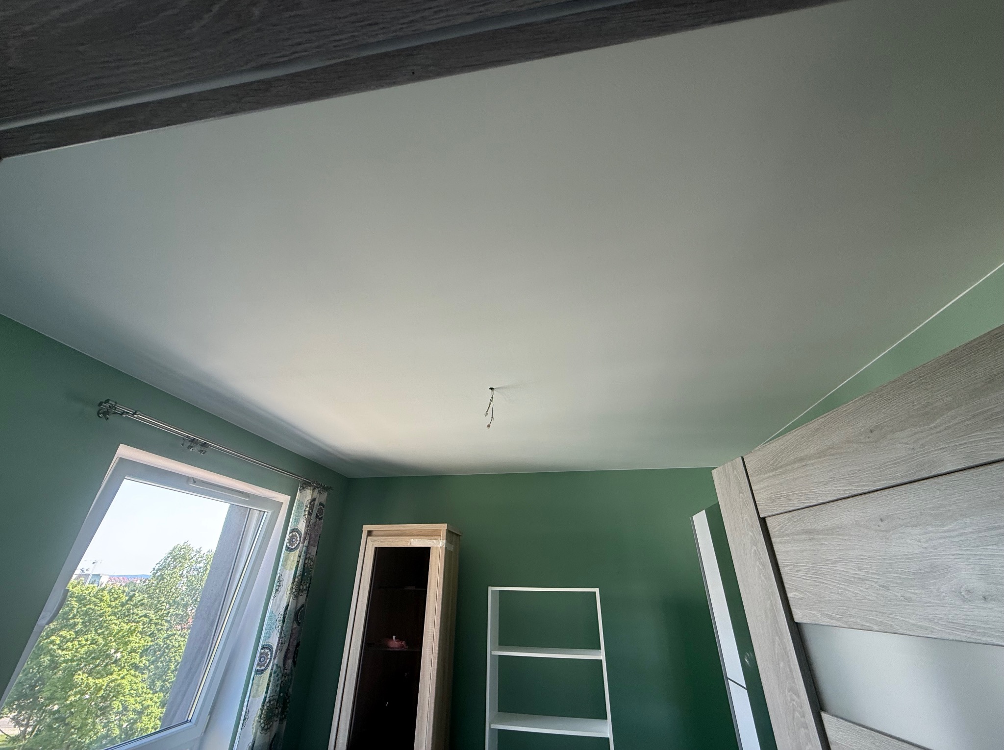 Pomieszczenie po malowaniu ścian na zielono i sufitu na biało. Widoczne okno dachowe, drzwi, regał i szafka. W suficie wystają kable.