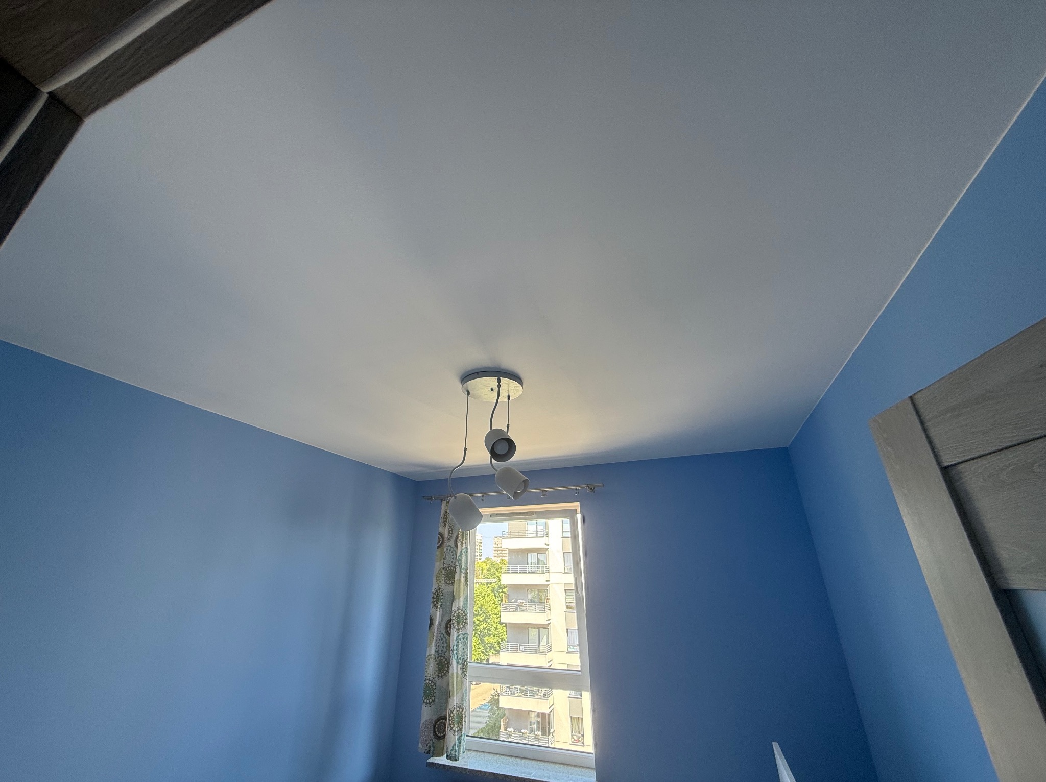 Błękitne ściany i biały sufit w pokoju z oknem. Widoczna lampa sufitowa, fragment drzwi i okno z widokiem na budynek.