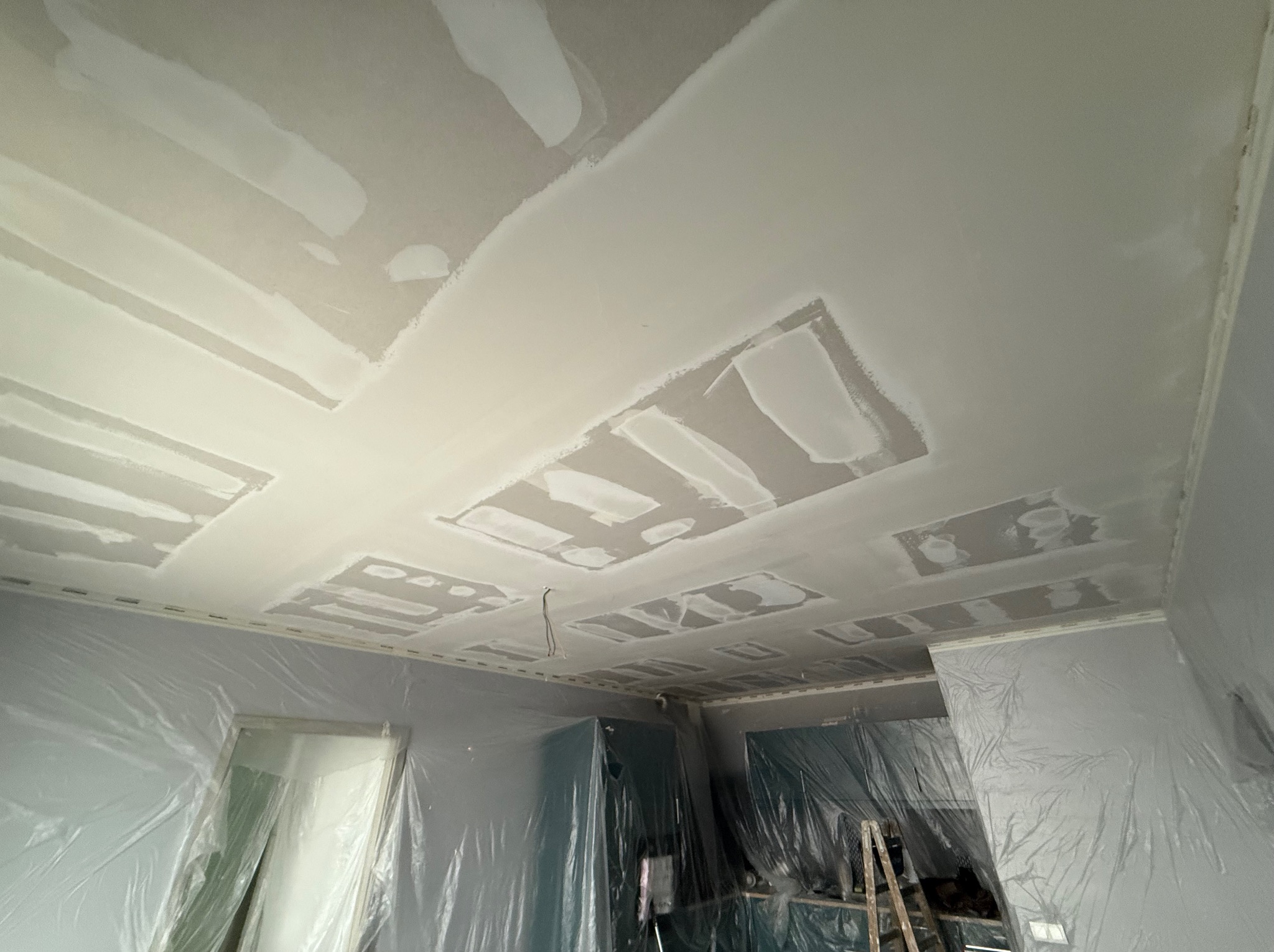 Sufit po szpachlowaniu z widocznymi łatami i zabezpieczonymi folią ścianami, przygotowany do dalszych prac wykończeniowych.