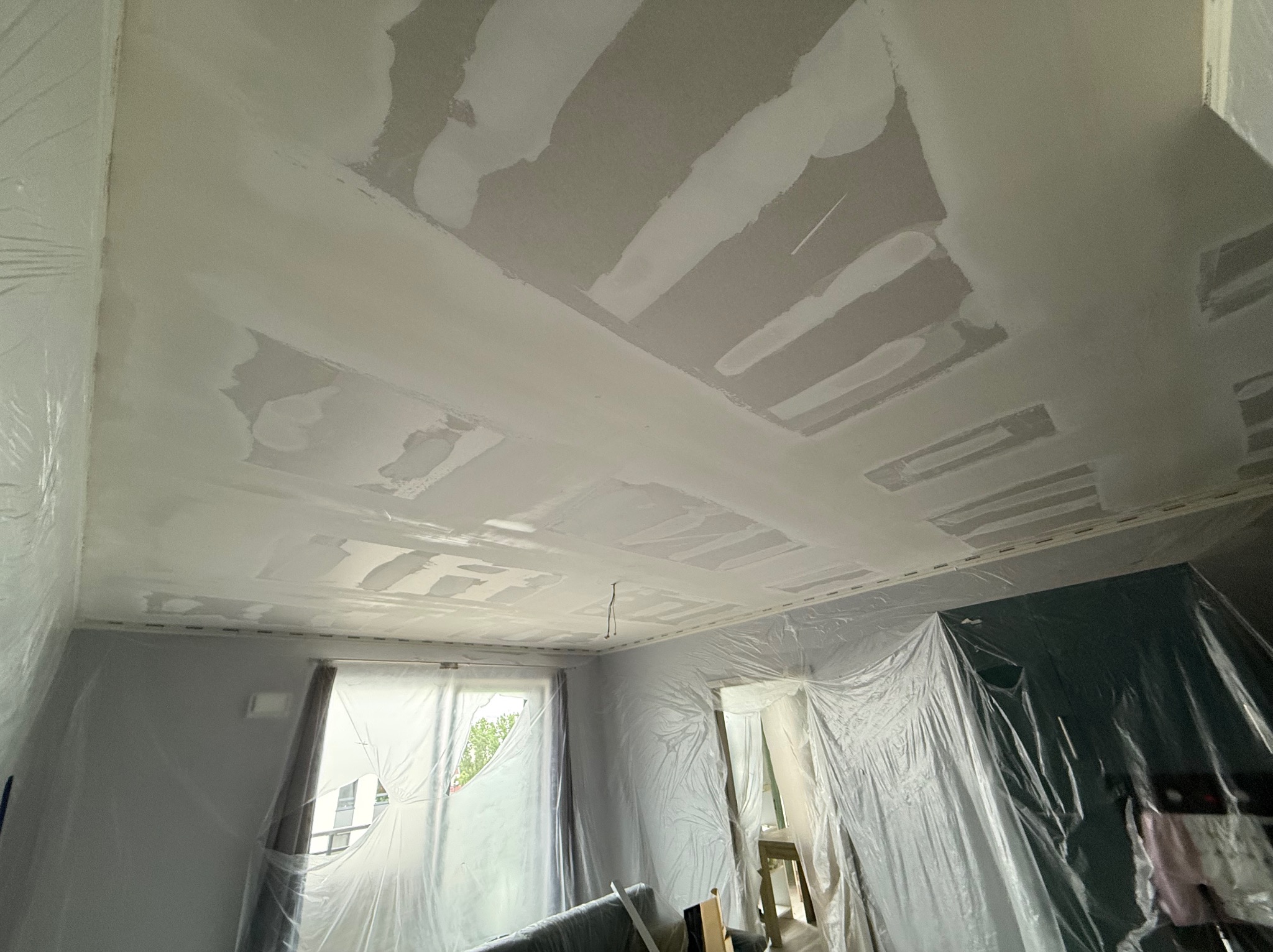 Pokój w trakcie szpachlowania sufitu i ścian, zabezpieczony folią malarską. Widoczne ślady szpachli na suficie, okno z widokiem na zieleń.