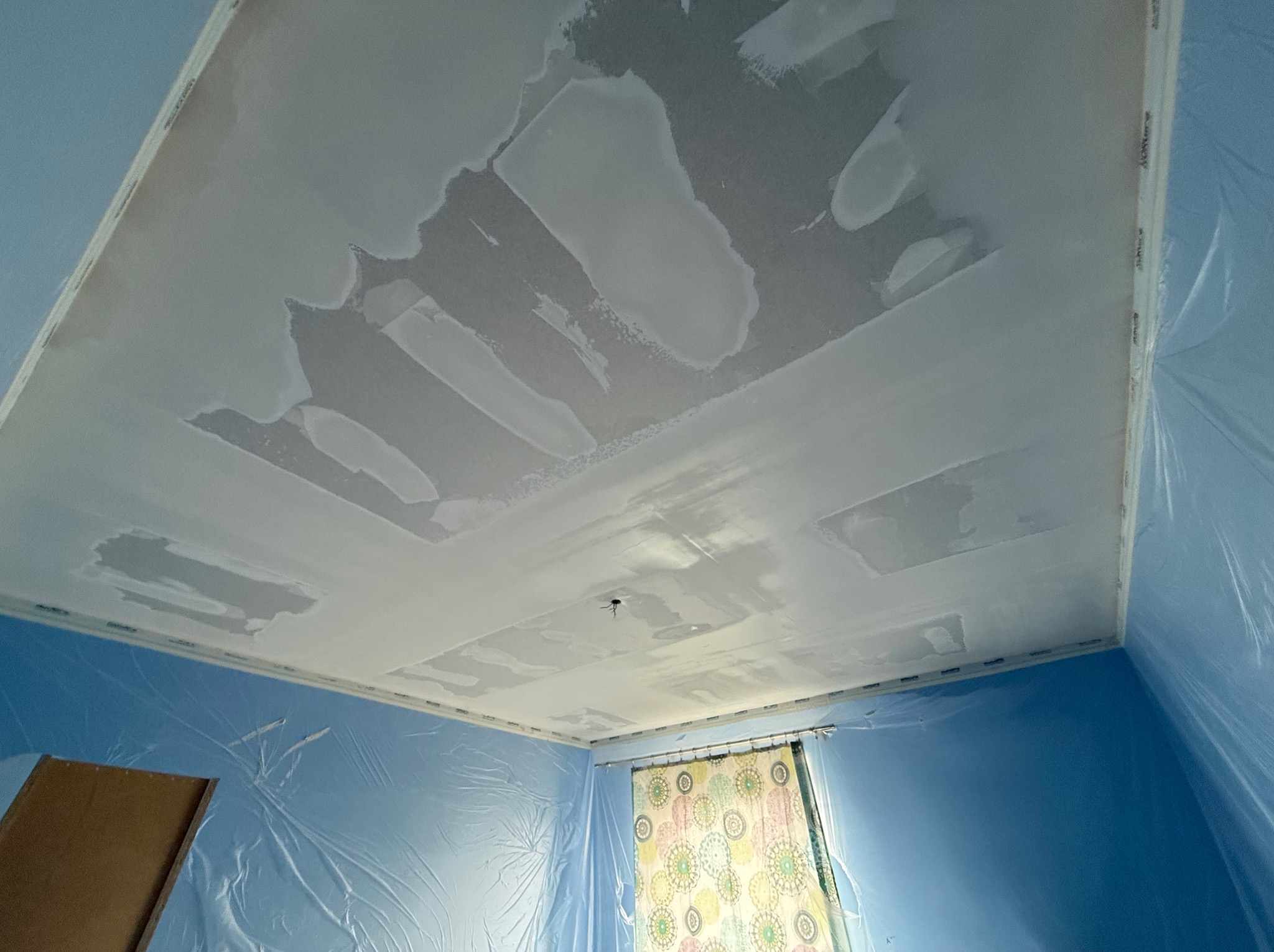 Nierównomiernie nałożona gładź na suficie z widocznymi łatami, zabezpieczona folią ściana w kolorze niebieskim i zasłonięte okno.